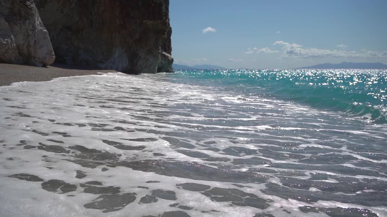 在阳光明媚的日子里，在巨石附近的海滩上，长长的海浪带着泡沫。视频素材