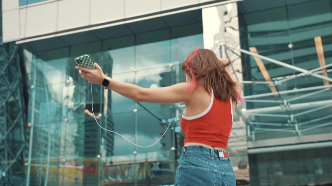 一个带着耳机拿着智能手机在城市街道上跳舞的时髦女人视频素材