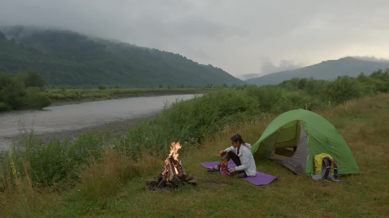 女人坐在山上的帐篷旁边，欣赏着河边的景色视频素材