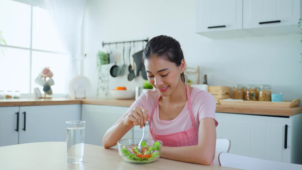 穿着围裙的亚洲美女在家里的厨房里吃着蔬菜沙拉。年轻美丽的女孩感到快乐，享受早餐与蔬菜健康的食物和清洁的水，以饮食的健康在家里。视频素材