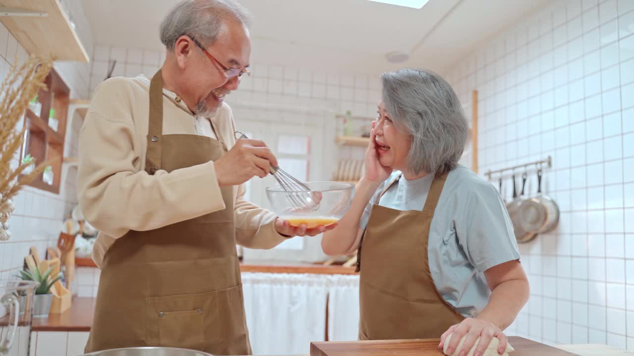 亚洲老夫妇站在厨房里，幸福地享受着退休生活。丈夫搅鸡蛋，妻子打面粉，玩粉。爱和家庭活动的关系。视频素材