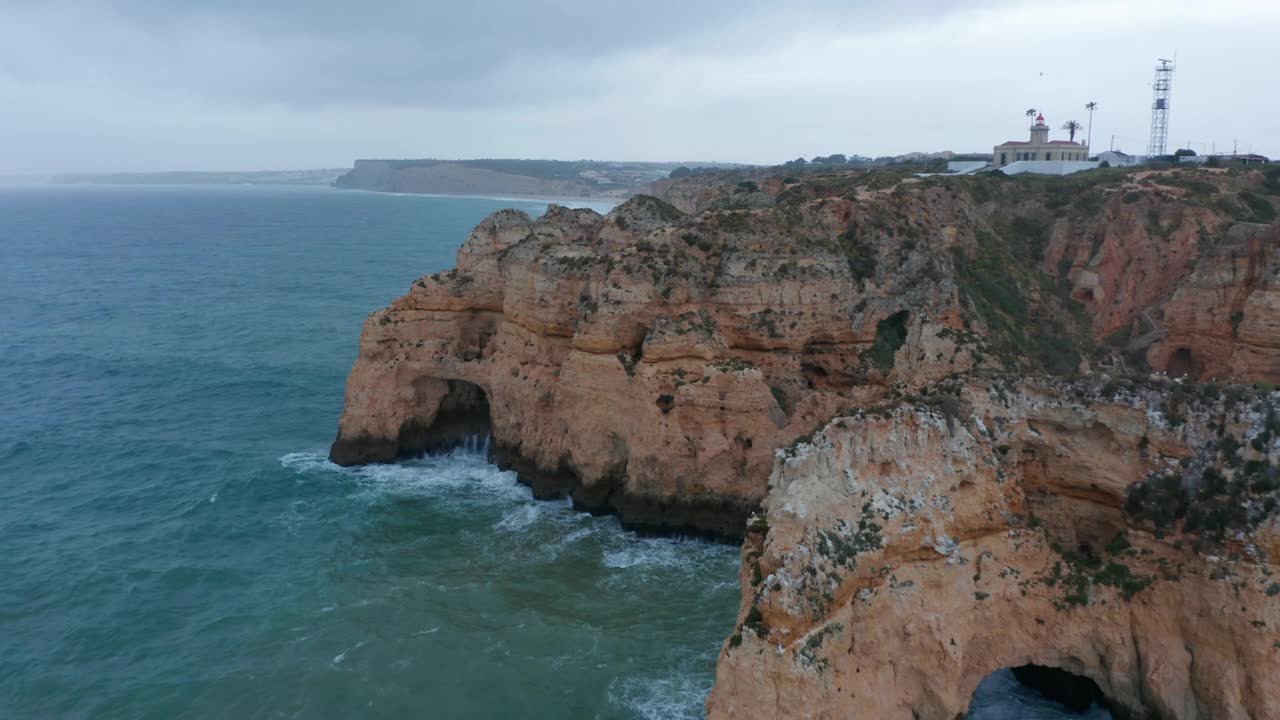 在多云的日子，在葡萄牙拉各斯的彩色岩石悬崖附近，沿着海岸与灯塔的背景飞行的无人机视频素材