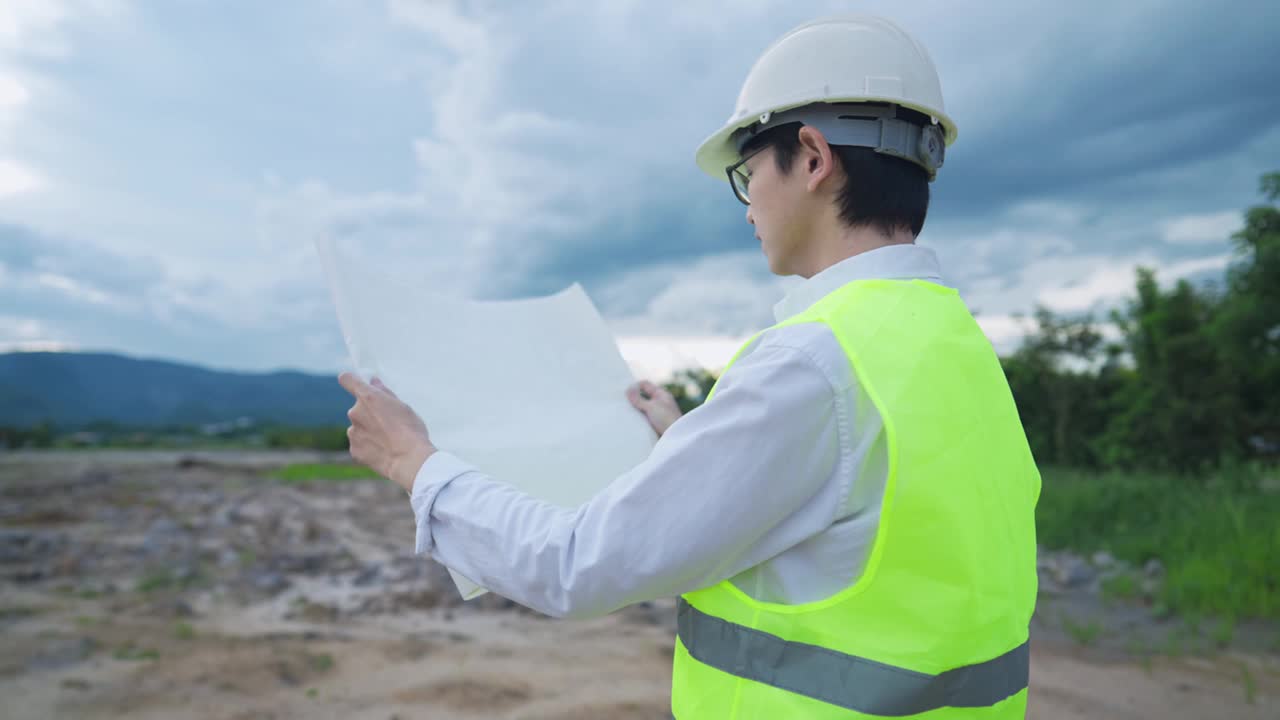 年轻的专业高级工人站在农村荒地上看建筑结构图纸，手里拿着设备开发计划，安全穿着工人，施工工人穿视频素材