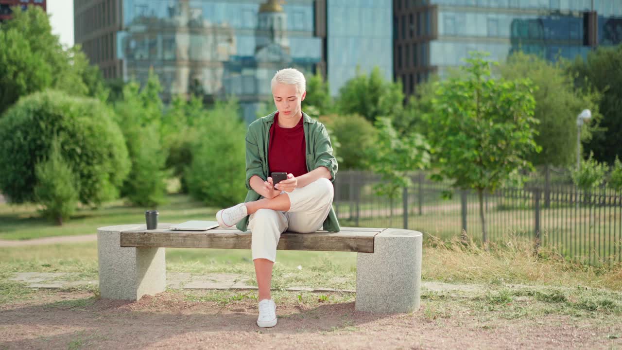 放大年轻的女商人与短金发打字短信和看着相机自信地坐在长凳上，笔记本电脑和外卖咖啡杯附近。女人用手机发短信视频素材