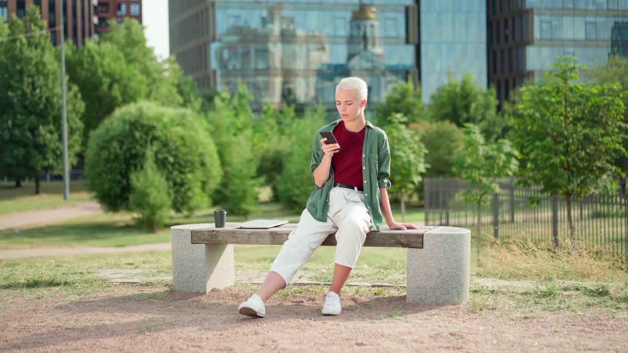 金发短发的女企业家或女大学生坐在公园的长椅上，一边用手机发短信，一边做着白日梦，旁边放着笔记本电脑和外卖咖啡杯视频素材