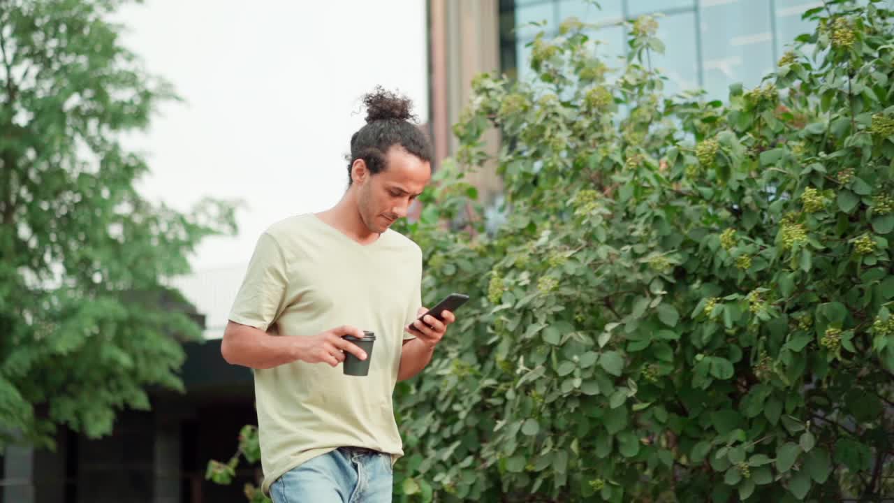 跟踪侧视慢镜头，年轻男子拿着外卖咖啡杯走在城市街道上，用手机发短信或上网，女子带着笔记本电脑经过视频素材