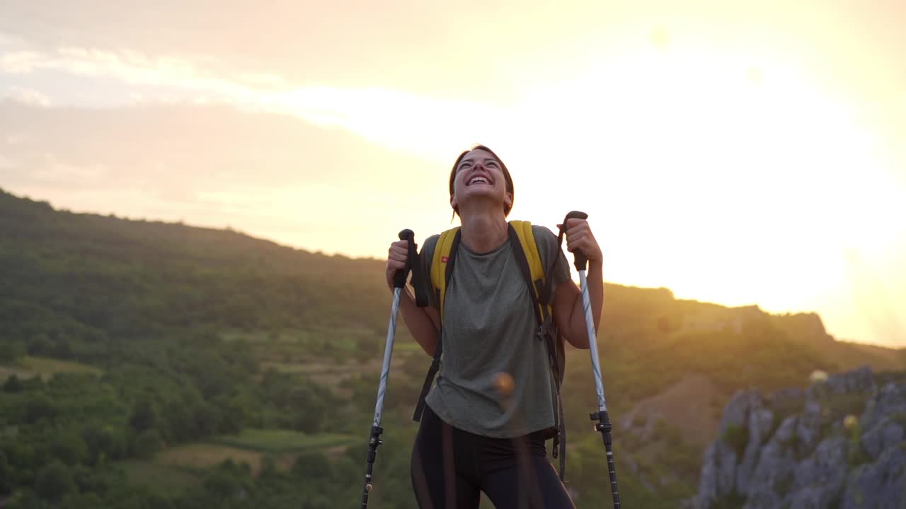 兴奋的年轻女性徒步旅行者，庆祝她成功地在宏伟的日落在她身后的山顶视频素材