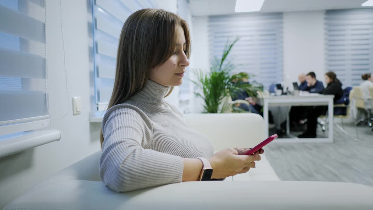 一个现代女孩在办公室的沙发上使用手机，滚动，打字，上网。应用的概念，信使视频素材