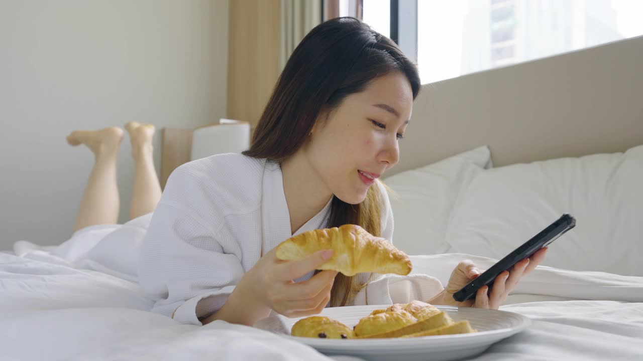 美丽的亚洲年轻女子穿着浴袍在卧室的床上吃早餐时使用智能手机视频素材