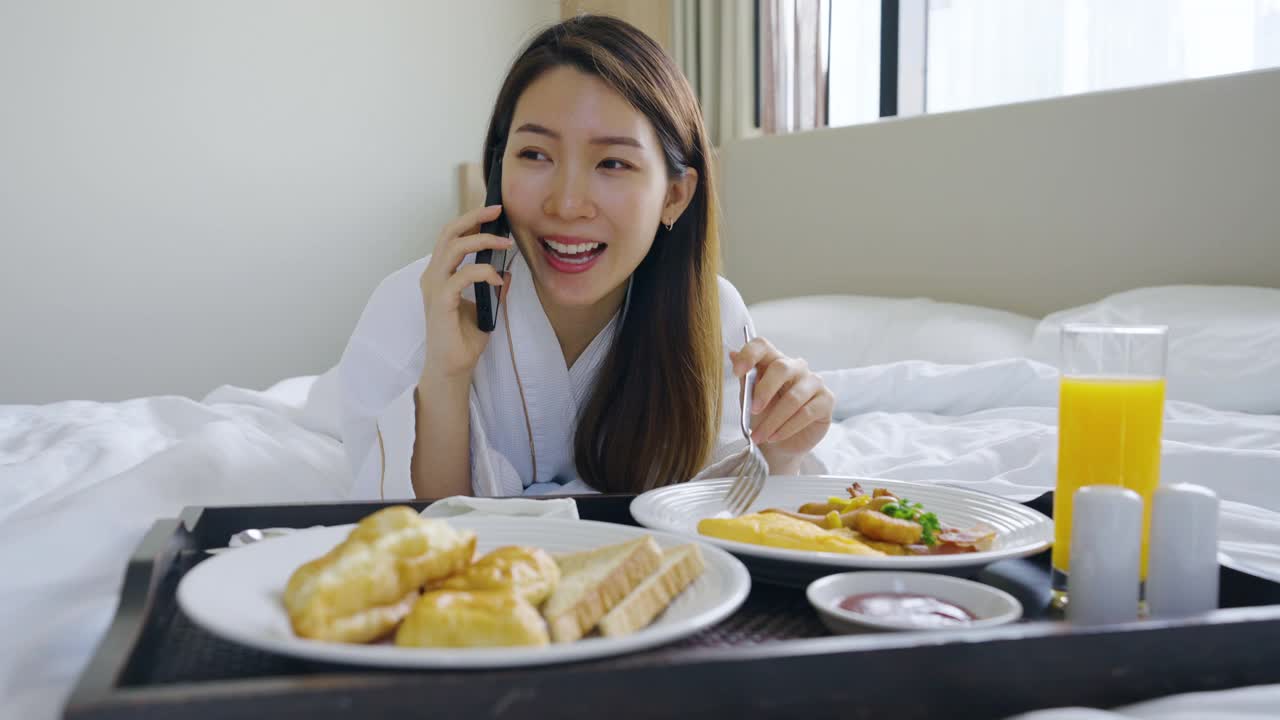 美丽的年轻亚洲女人穿着浴袍在卧室的床上吃早餐的时候讲电话视频素材