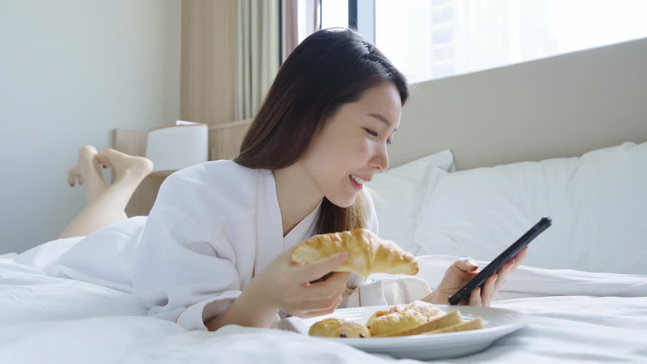 美丽的亚洲年轻女子穿着浴袍在卧室的床上吃早餐时使用智能手机视频素材