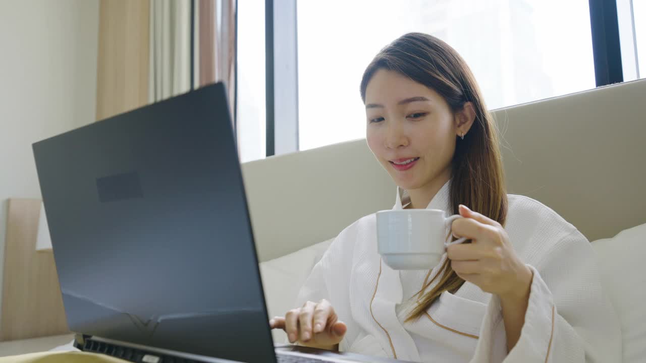 亚洲女人的肖像穿着浴衣使用笔记本电脑，在卧室的床上喝早上的咖啡视频素材