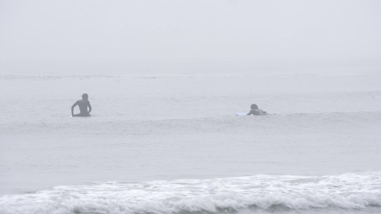 从漂浮在海上冲浪板上的冲浪者的中等角度降至两名携带冲浪板出海的人。视频素材