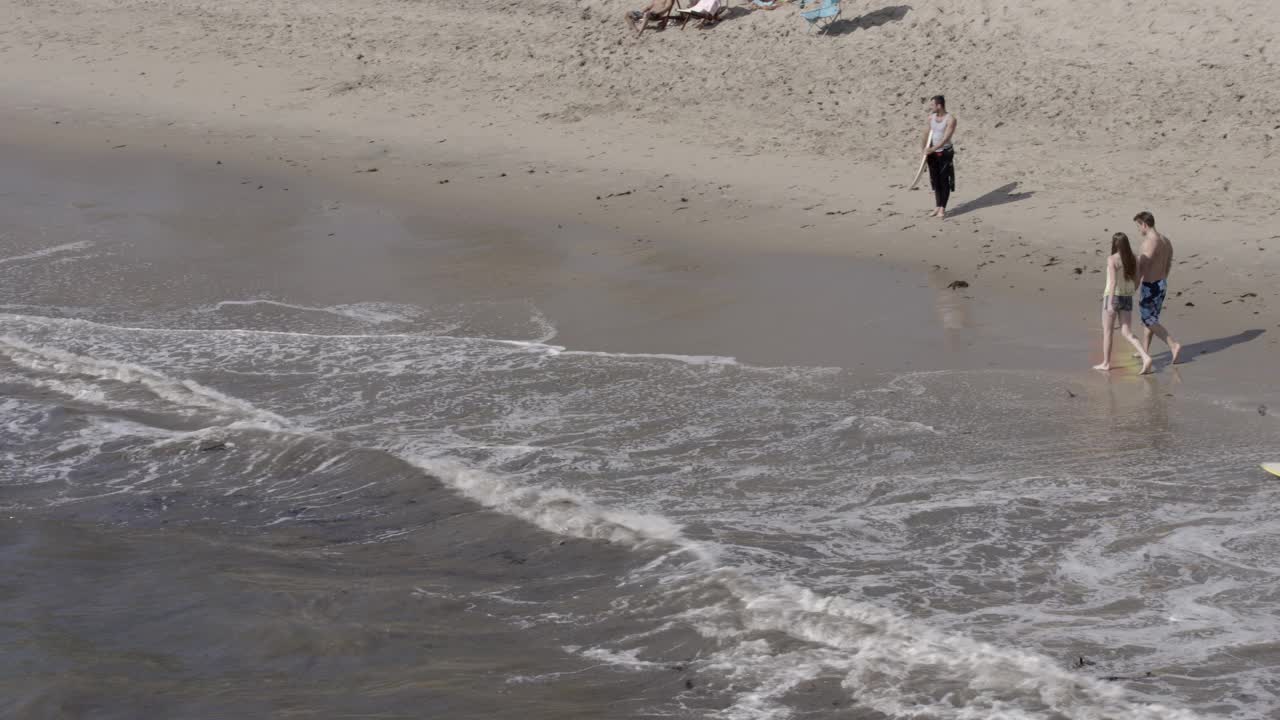 从在海浪中冲浪的人到海滩小屋。海滩上可以看到穿泳衣的人。海滨别墅前可见的帆船。视频素材