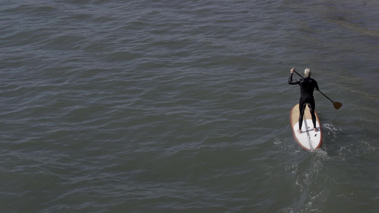 从穿着潜水服在海边玩冲浪板的人到海滩别墅。海滩上可以看到穿泳衣的人。海滨别墅前可见的帆船。视频素材