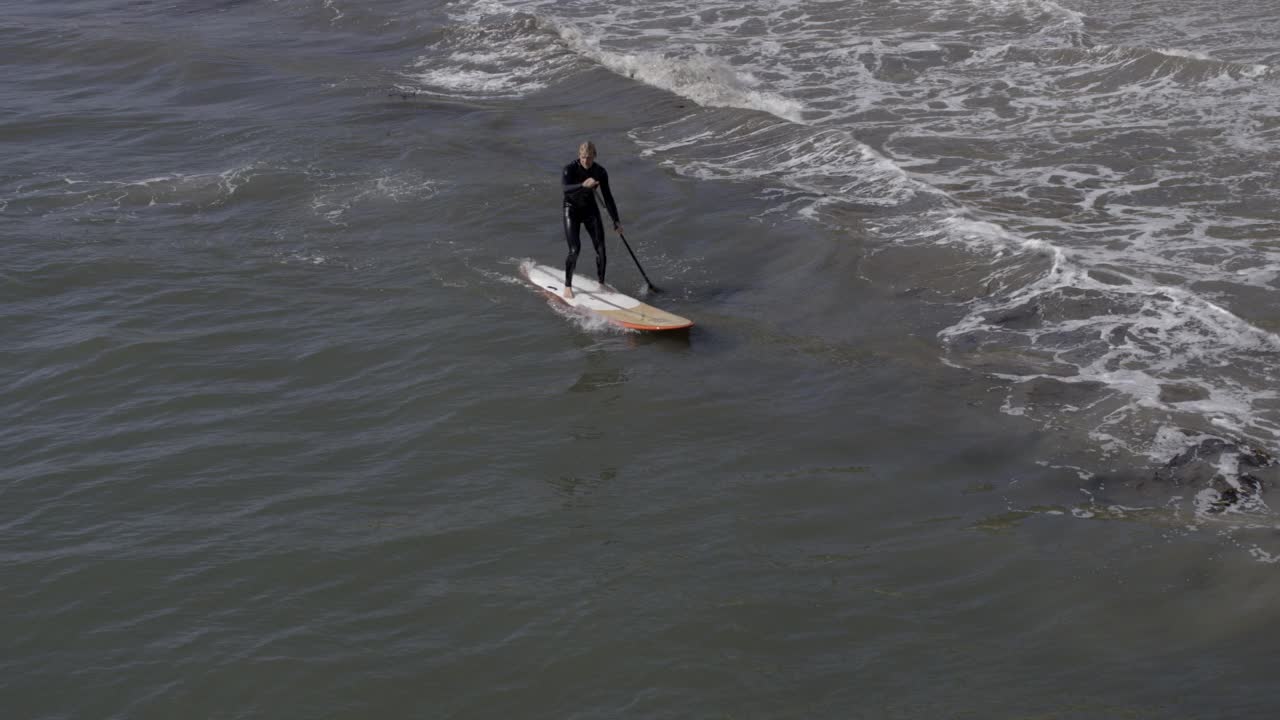 从穿着潜水服在海边玩冲浪板的人到海滩别墅。海滩上可以看到穿泳衣的人。海滨别墅前可见的帆船。视频素材