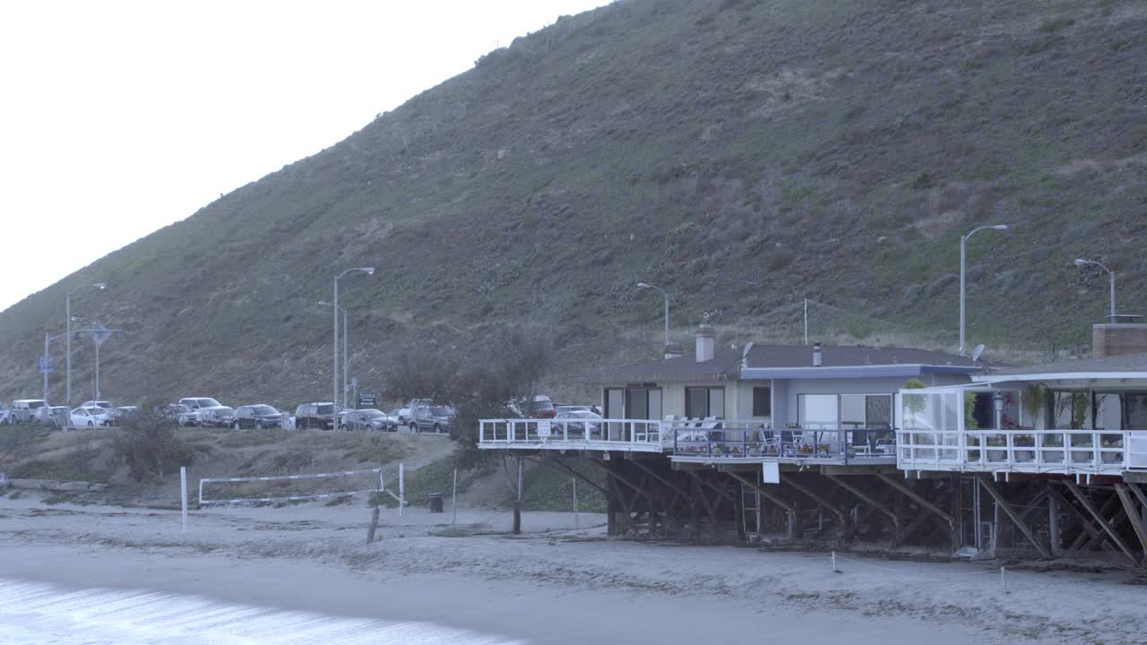 潘从左到右穿过海滩上的房子。太平洋沿岸公路上停着的汽车随处可见。排球网、沙滩、房子前的帆船。冲浪者拿着冲浪板从右到左走过。视频素材