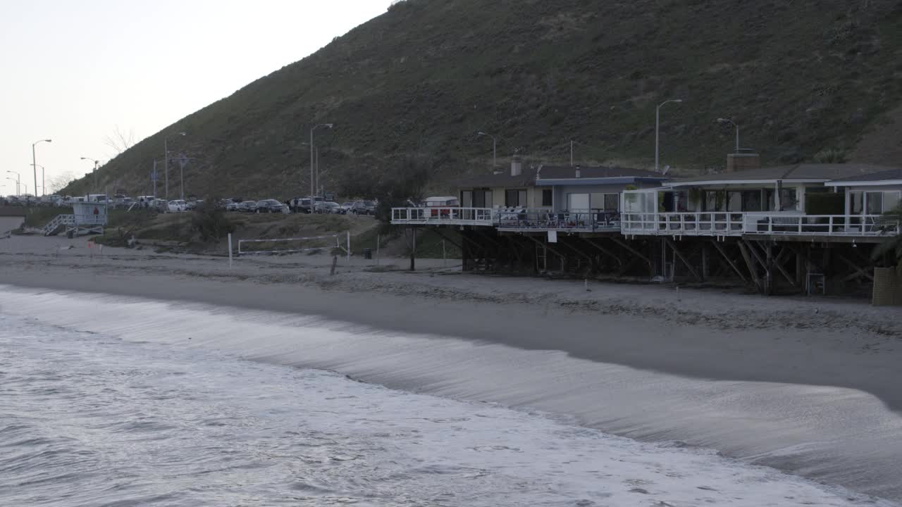 潘从左到右穿过海滩上的房子。太平洋沿岸公路上停着的汽车随处可见。排球网、沙滩、房子前的帆船。bg中可见救生员塔。视频素材