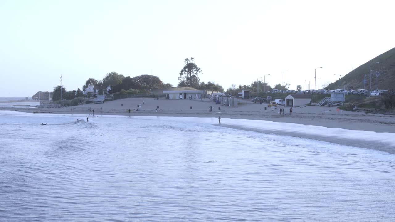 潘从左到右的广角从冲浪的人在海洋中冲浪到排的海滩房子。海滨别墅前可见的帆船。救生员塔、沙滩和排球网随处可见。视频素材