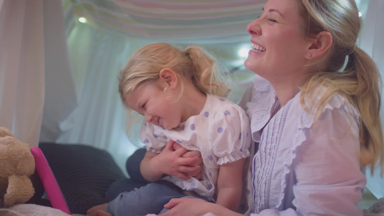 母亲和年幼的女儿在孩子的卧室里的自制营地里看电子平板电脑视频素材