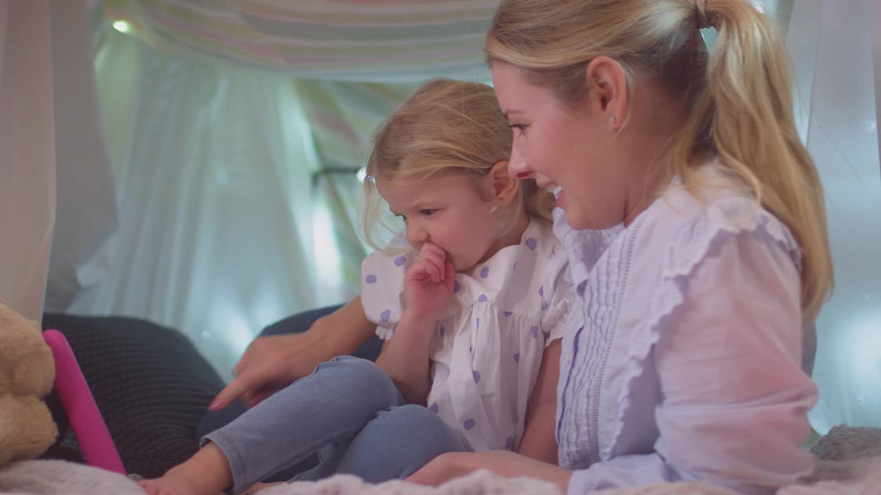 母亲和年幼的女儿在孩子的卧室里的自制营地里看电子平板电脑视频素材