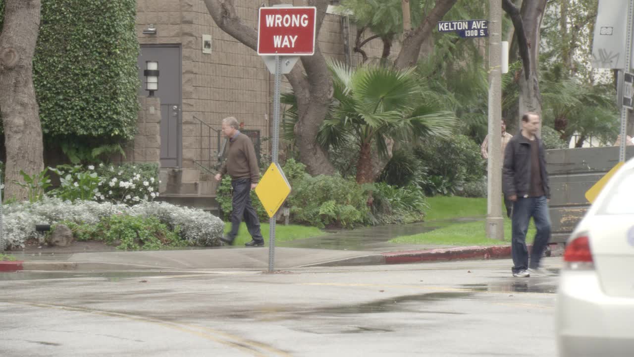 从右到左中等角度观察雨中人行道上的行人。一个男人带着伞。汽车停在街上。公寓建筑部分可见。罗彻斯特大街10966号的建筑遮阳篷。视频素材