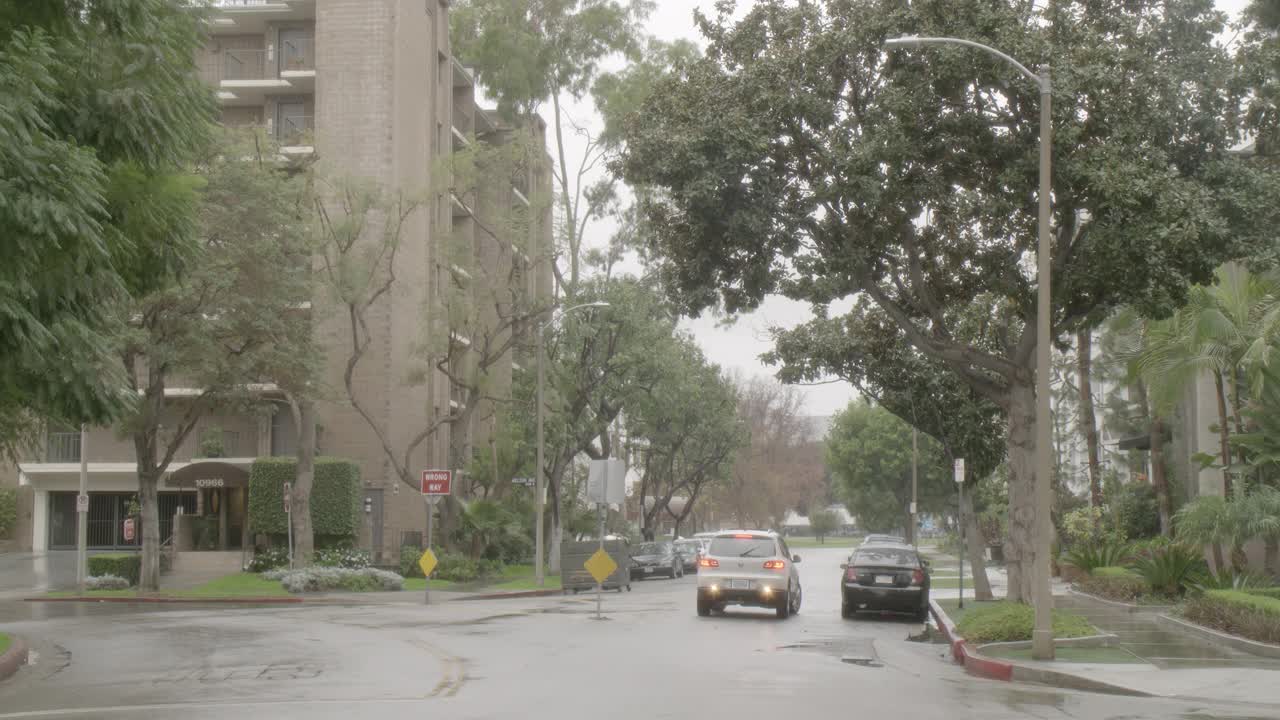 雨中城市街道的中等角度。可见多层公寓楼。汽车驶过，其他人停在路边。视频素材