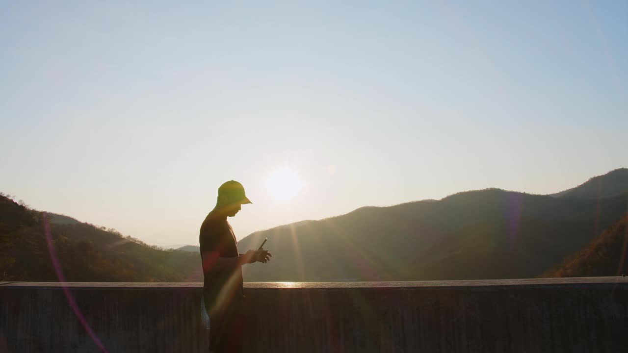 嬉皮士徒步旅行者英俊的男人站在山上，用智能手机放松，看着美丽的自然，看着夕阳的背景视频素材