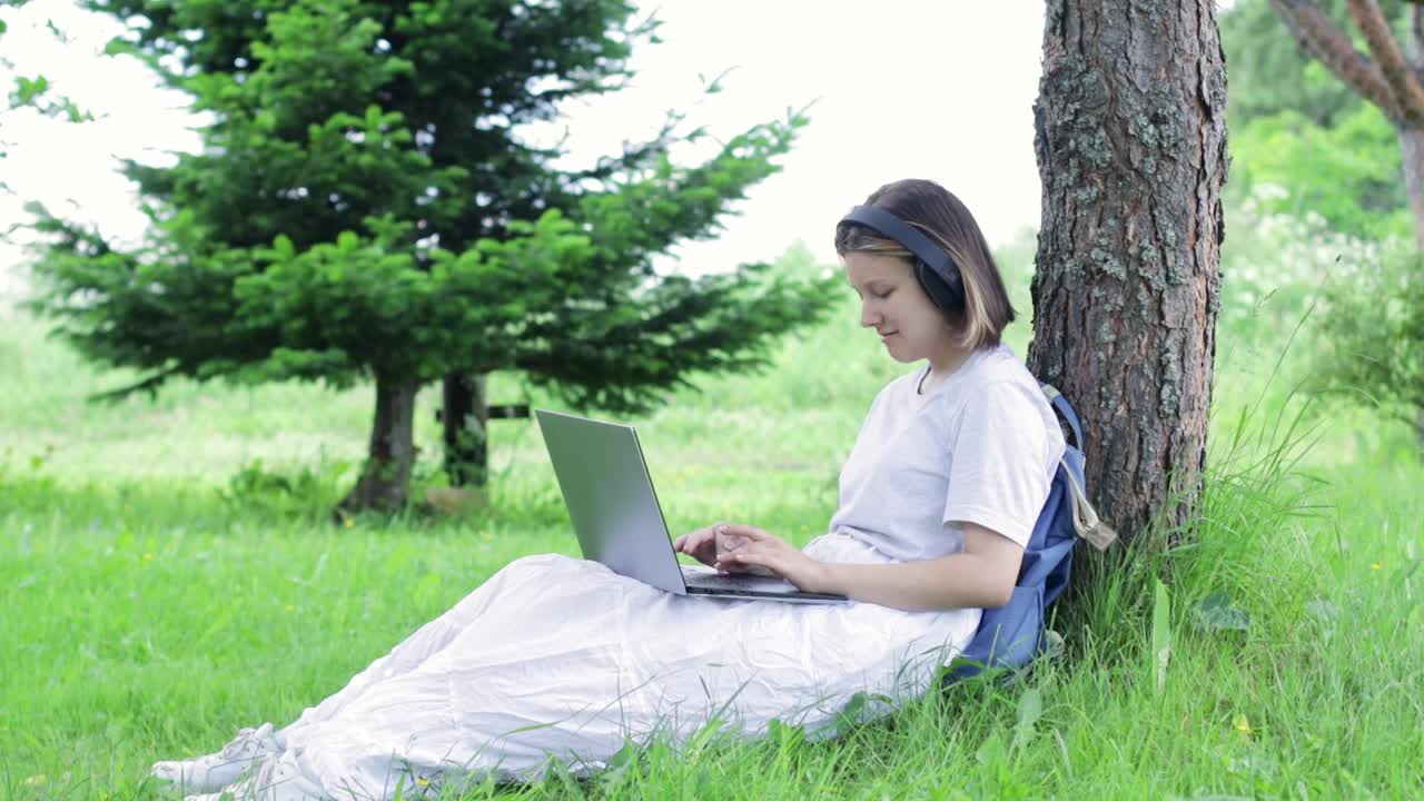 十几岁的女孩坐在公园的一棵树旁，用笔记本电脑打字视频素材