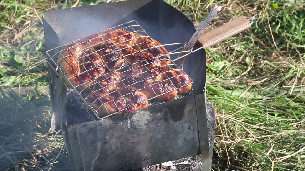 烤架烤香肠。烟和燃烧的煤块一起从烤架上冒了出来。在绿草如茵的草地上野餐。特写镜头。视频素材