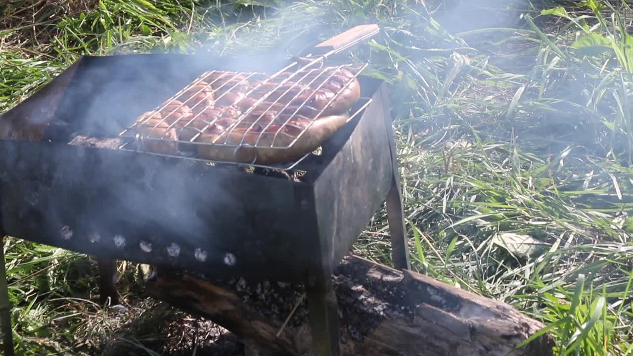 烧烤架与棕色香肠。躺在烤架上，还有余烬。在绿草如茵的草地上。野餐。特写镜头视频素材