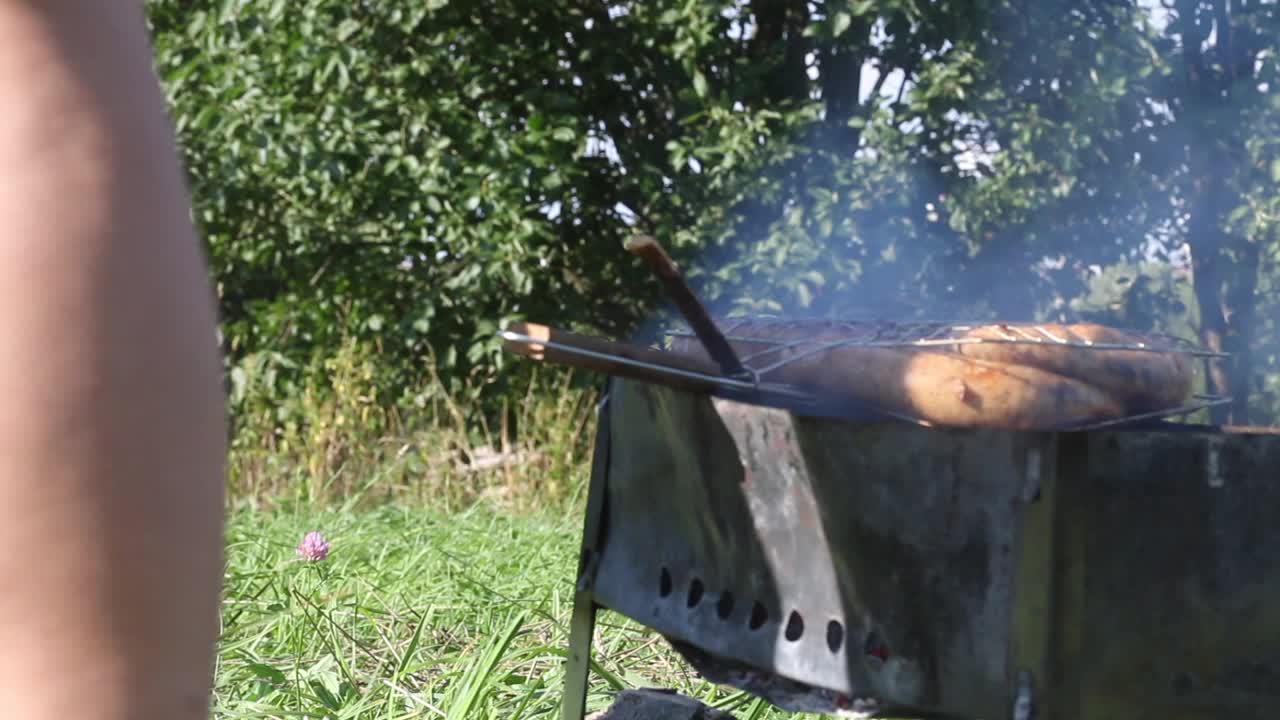 这个人把烤香肠翻过来。在木炭烤架上烹饪在绿草如茵的草地上野餐。特写镜头视频素材