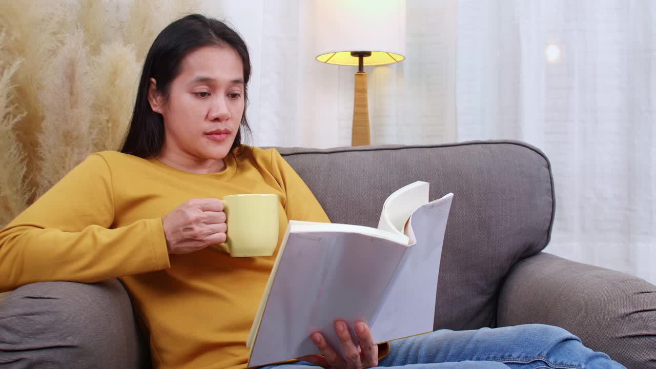 年轻女子在舒适的客厅里看书。女人坐在或躺在家里的沙发上看书。女性在周末的空闲时间在室内舒适的沙发上放松视频素材