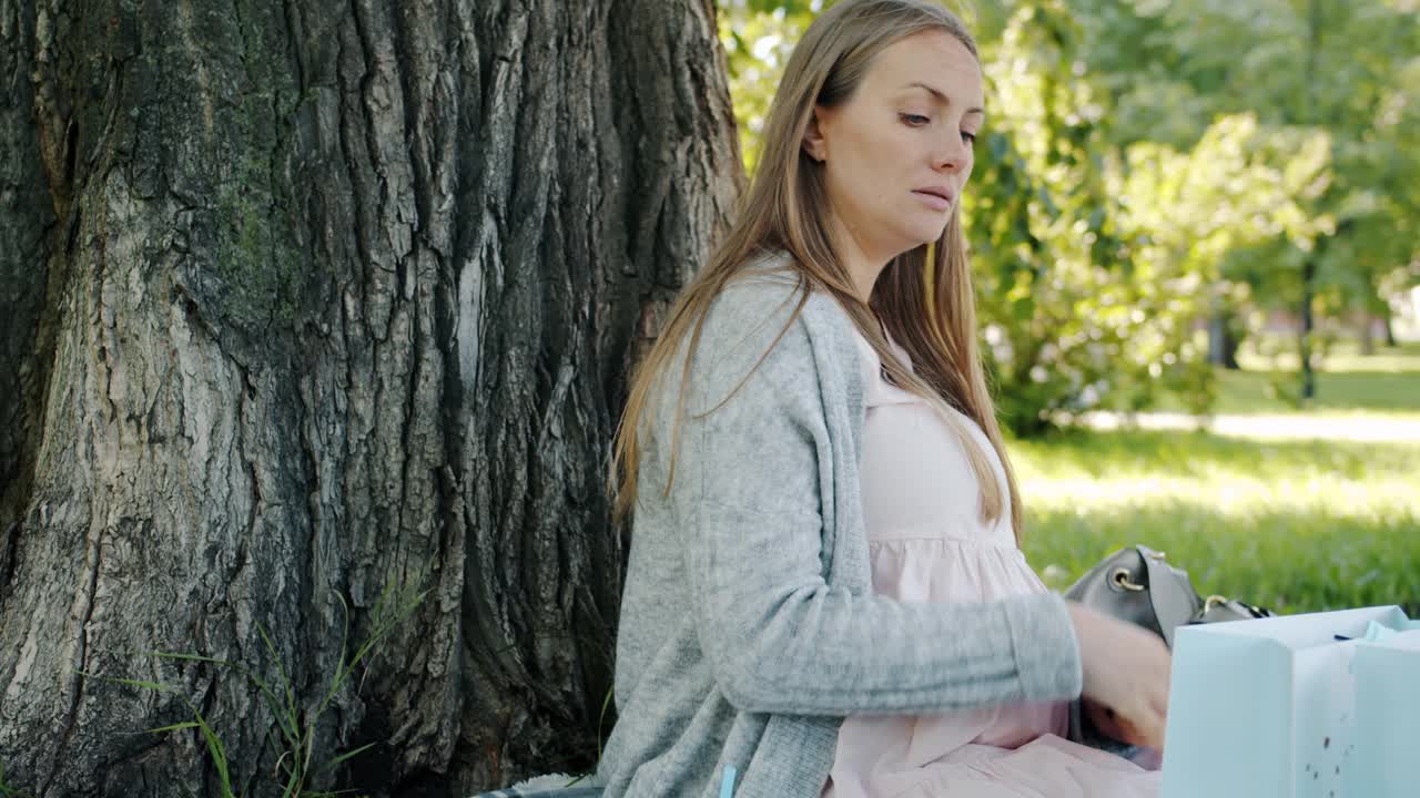 快乐的孕妇看着新的婴儿衣服，打开购物袋坐在公园的草坪上视频素材