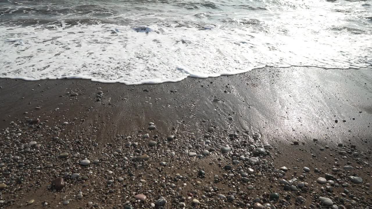 高角度的观点，泡沫的海洋海水滚动的沙滩小岩石在阳光户外。日落时分，干净的地中海海浪在阳光下飞溅。风景如画的海景。视频素材