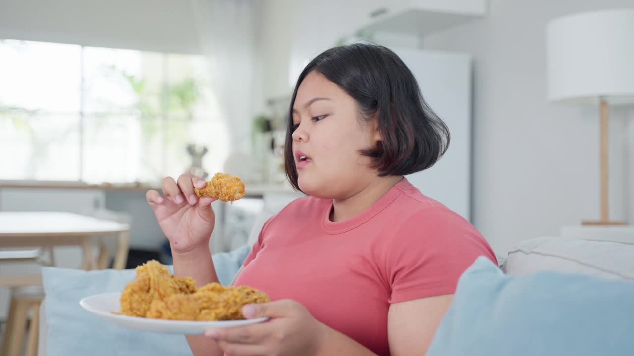 亚洲胖女人的肖像在家里吃不健康的食物。漂亮的大码女孩感到饥饿和快乐，在客厅的沙发上咬炸鸡，微笑，看着摄像机。视频素材