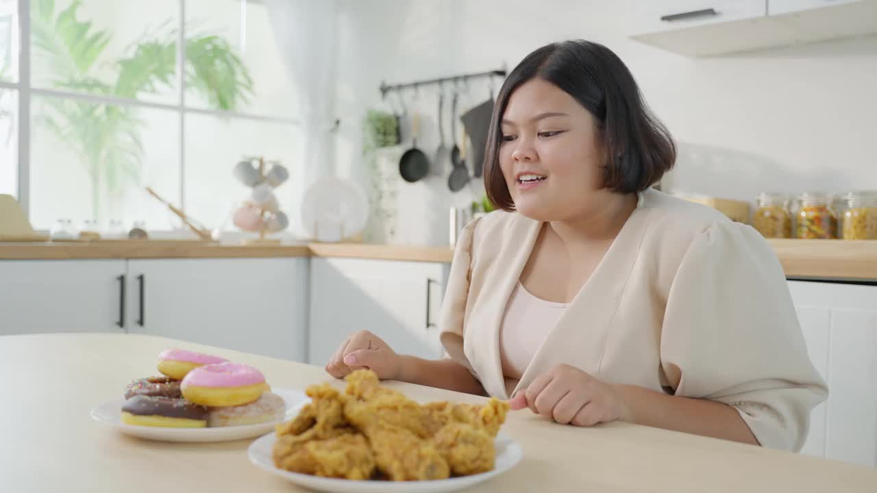 亚洲年轻的胖女人为盘子里的不健康食物感到高兴。诱人的美丽的加码女孩感到饥饿，准备吃炸鸡和甜甜甜圈在家里的餐桌上。视频素材