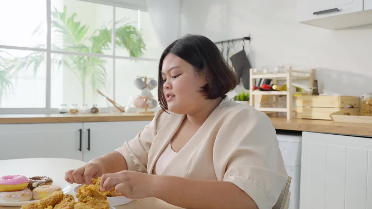 亚洲年轻的胖女人在家厨房里吃不健康的食物。迷人的美丽的大码女孩感到饥饿和快乐的咬炸鸡和甜甜甜圈在家里的餐桌上。视频素材