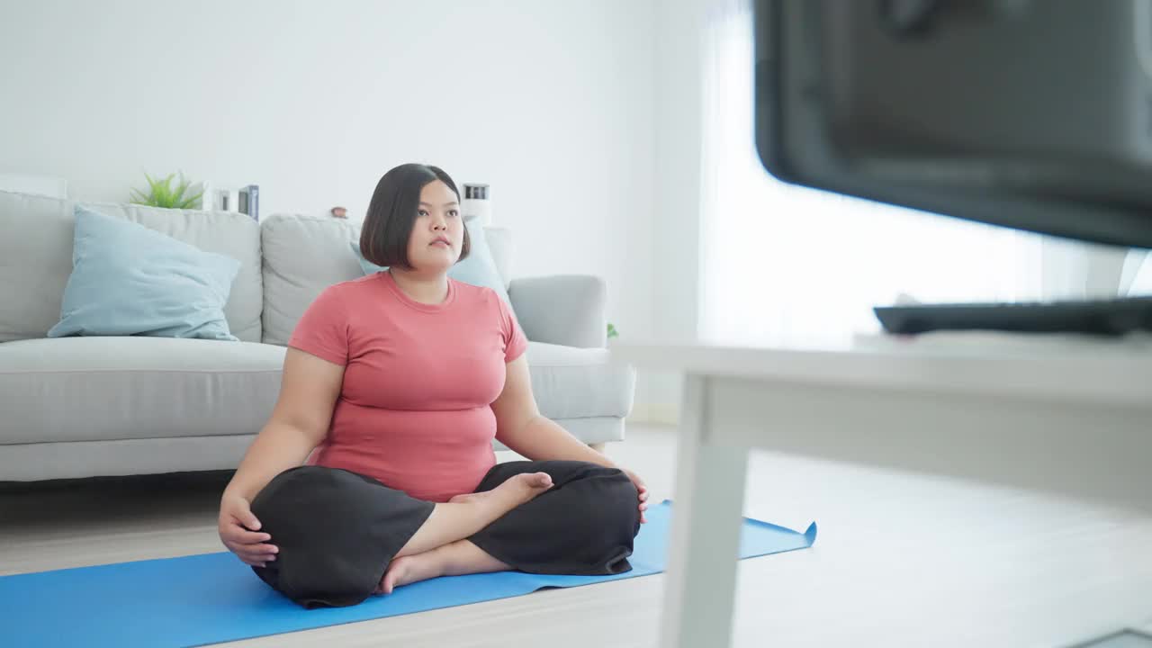 亚洲年轻的特大号女人在家里的客厅里做瑜伽练习。吸引人的大码女孩锻炼遵循普拉提指导视频从在线教练在电视上，在家里减肥。视频素材