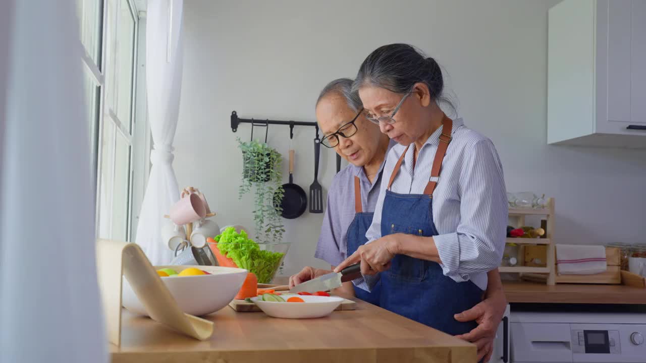 爱亚洲的老夫妇穿着围裙在厨房做饭。英俊强壮的老爷爷奶奶戴着眼镜，在家享受退休生活活动。家庭关系的概念。视频素材