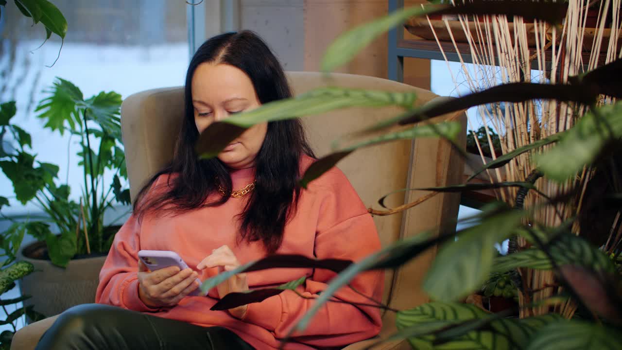 悠闲的女人在室内花园的软椅上使用手机。一名成年女性坐在橙色花圃里舒适的椅子上看智能手机。视频素材