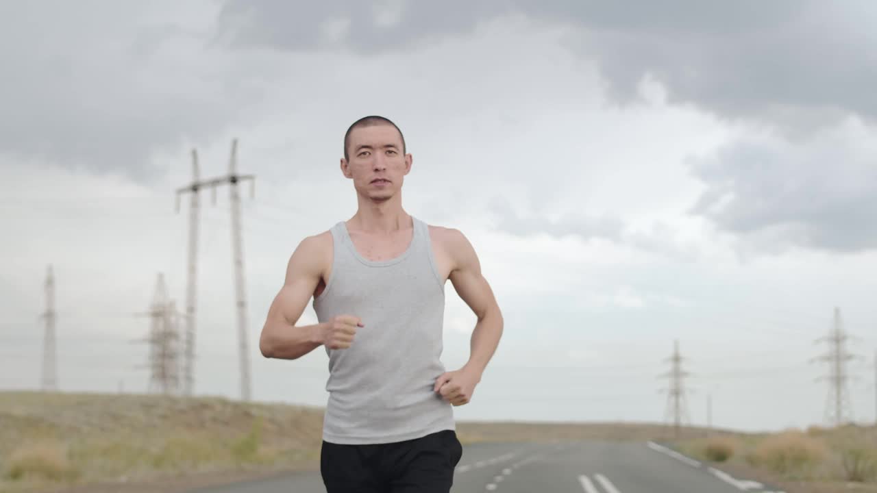 亚洲男人穿着运动服在路上跑，多云的天气，前面的景色。一名男子穿着草原T恤在高速公路上奔跑。体育的概念视频素材