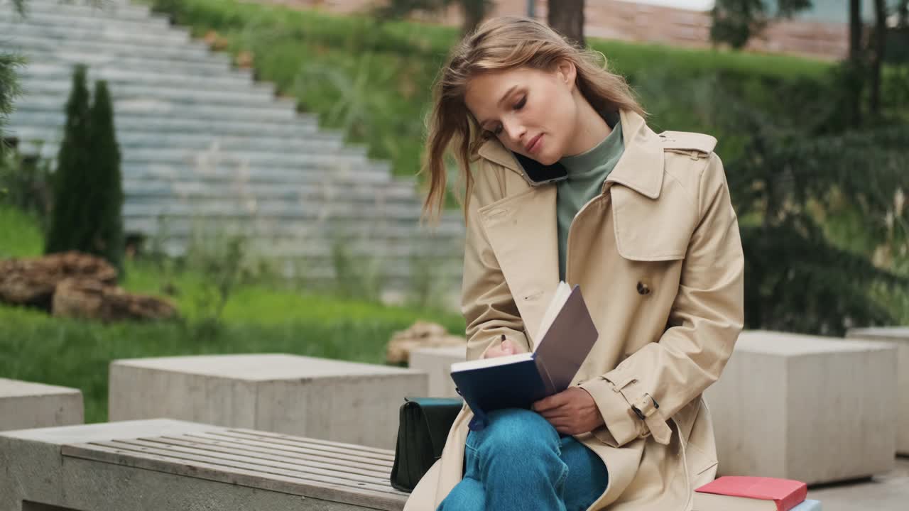 漂亮的女学生穿着风衣，在笔记本上做笔记，和朋友在户外打电话。金发女学生在公园的长椅上学习视频素材