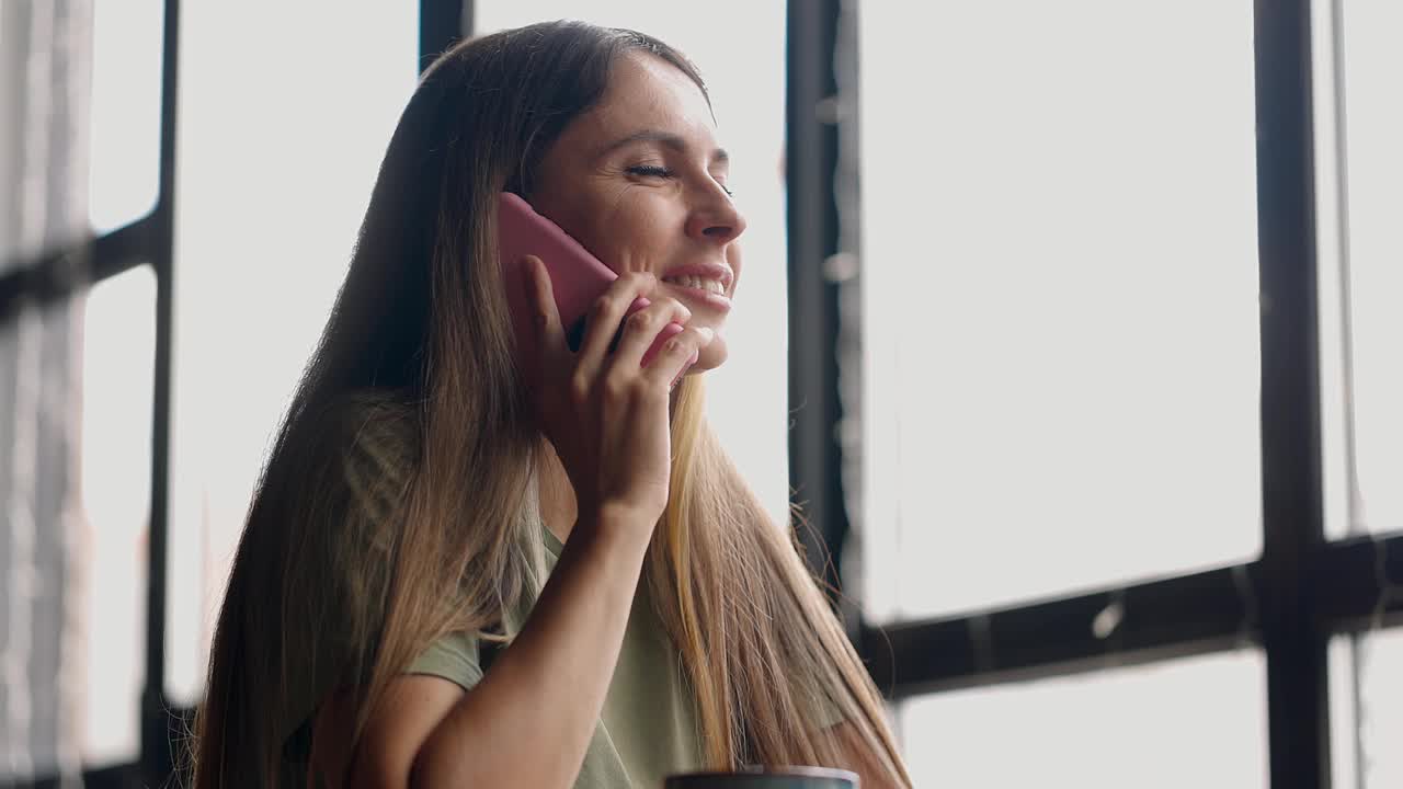 一个漂亮的女人坐在咖啡馆里，用手机聊天，微笑着看着窗外视频素材