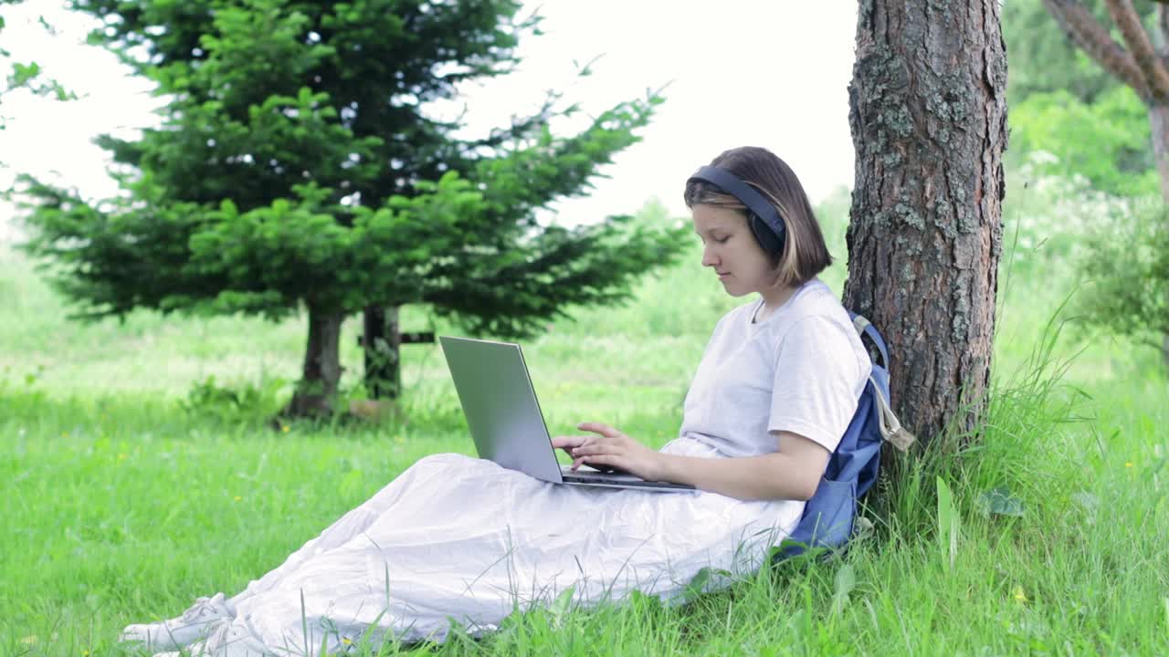 女孩坐在公园的一棵树旁，用笔记本电脑打字视频素材