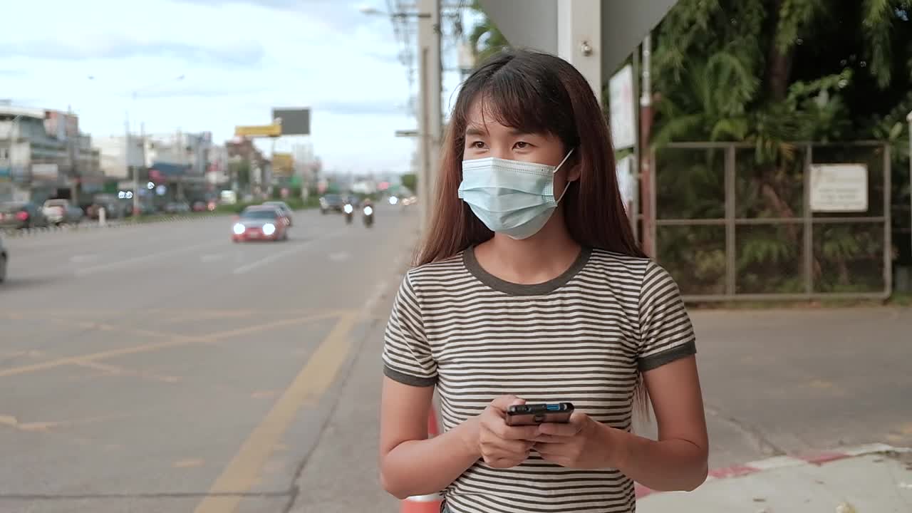 亚洲女子在城市购物街上唱着智能手机，戴着防止空气污染和新冠肺炎的口罩，走路和使用智能手机视频素材