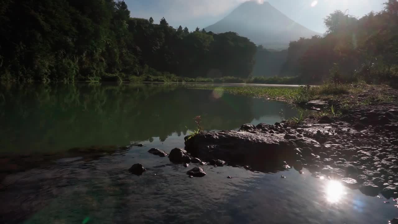 背景中出现了一个表面平静、清澈的湖水和喷出一股烟雾的火山。湖外长满了绿树。视频素材