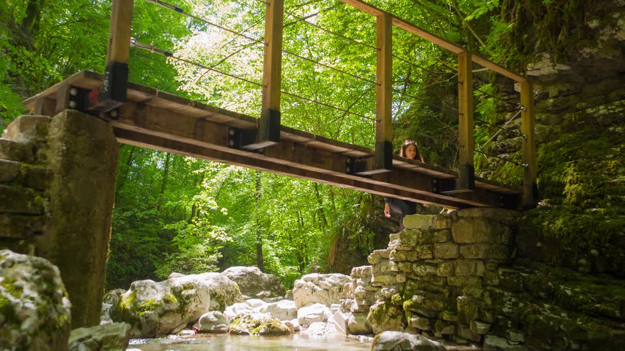 一名女游客在阳光明媚的日子徒步穿越山溪峡谷上的木桥视频素材