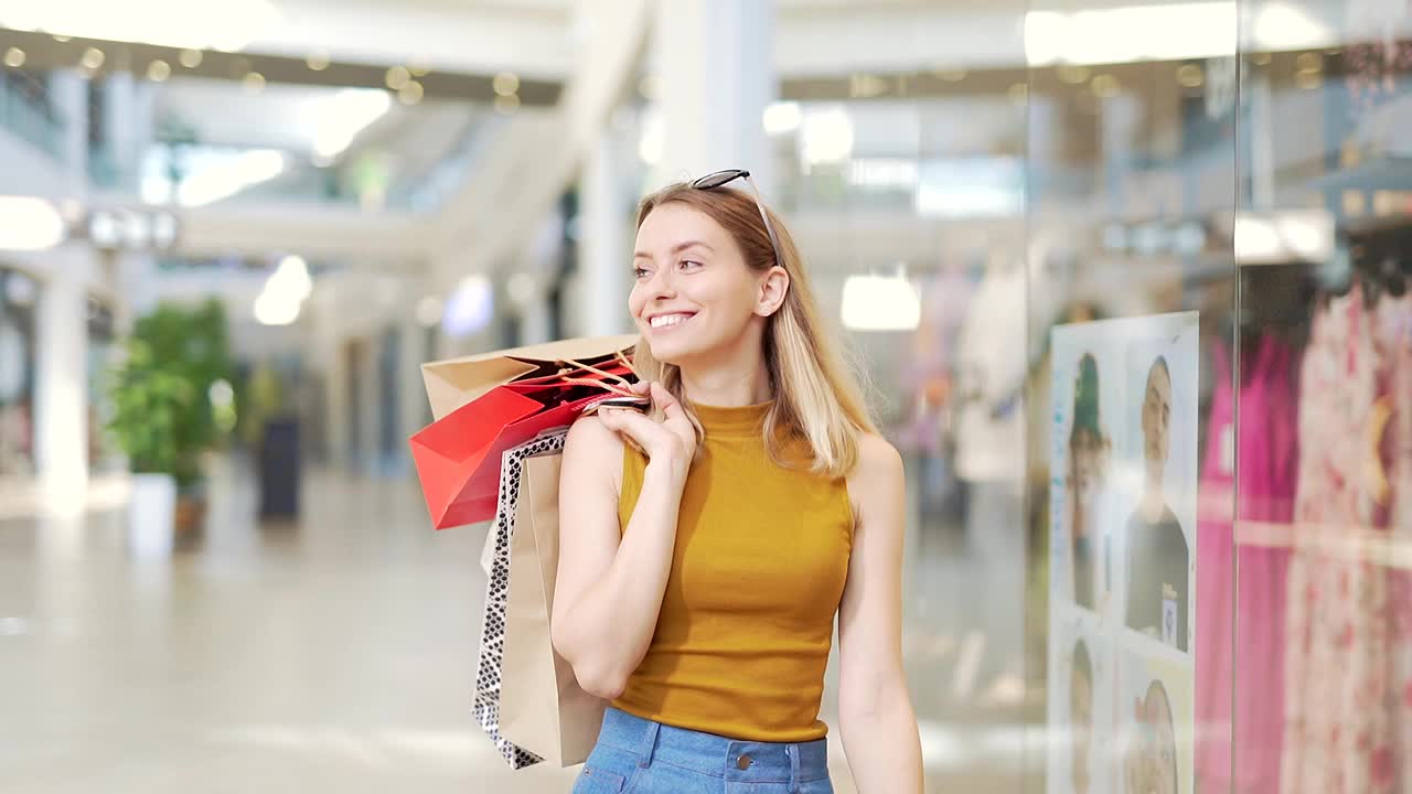 年轻快乐的金发美女花闲暇时间在大的现代购物中心。兴高采烈的女士提着五颜六色的购物袋视频素材