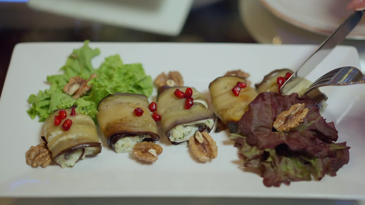 特写茄子卷，白色的盘子上有核桃。烹饪格鲁吉亚传统菜肴，女性的手用刀叉切割食物。不知名的客户品尝餐点。视频素材